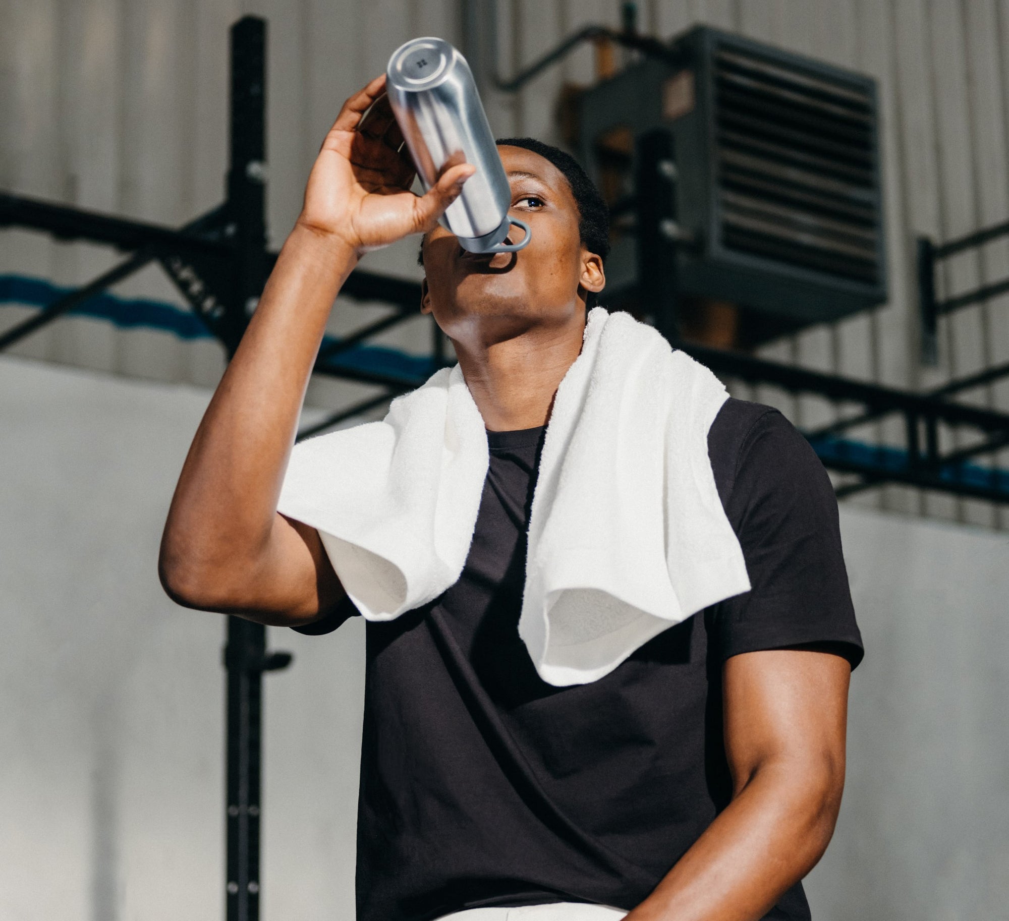 A man is sitting in a gym, with a towel hanging around his neck, drinking out of a waterdrop® Ultralight Steel bottle